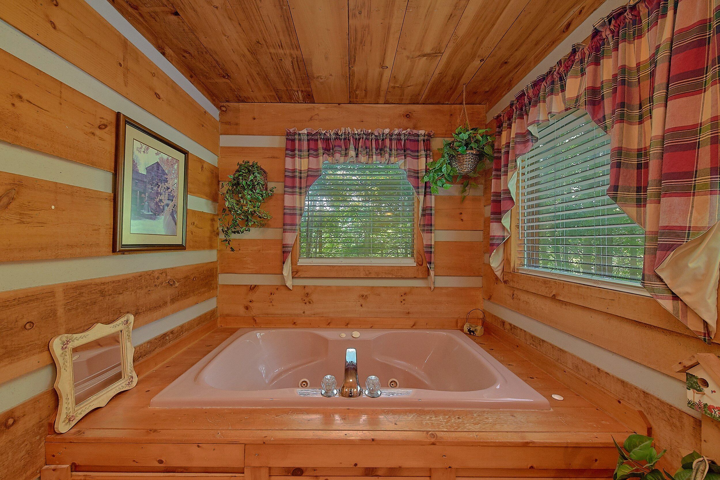A wooden cabin interior with a large bathtub, a framed picture on the wall, and a window with curtains.