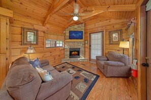 A cozy log cabin interior with a fireplace, wooden walls, a ceiling fan, and a television. - Camping Under the Stars Cabin Rental (Wears Valley)