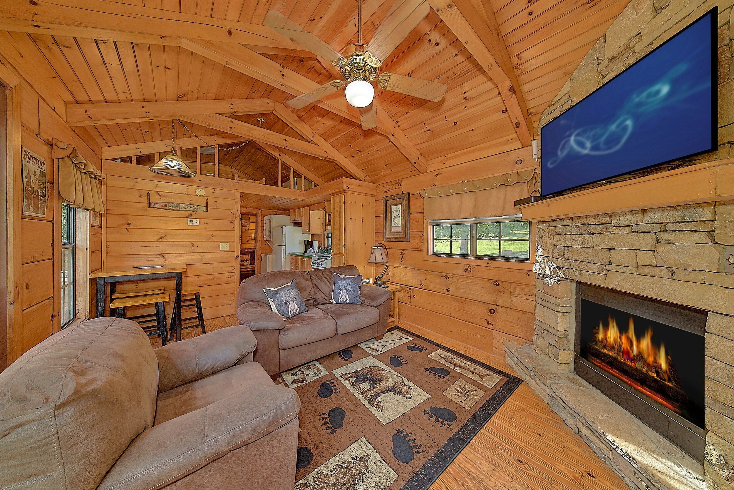 A log cabin interior with a fireplace, a television mounted on the wall, and a cozy seating area with a rug.