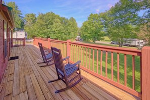 A wooden deck with rocking chairs and a red railing overlooking a grassy area with trees and a parked RV. - Camping Under the Stars Cabin Rental (Wears Valley)