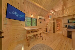 A wooden cabin interior with a dining area, kitchen, and a large window offering a view of the forest. - A Camper's Delight | Pet-Friendly Tiny Home w/ Fire Pit (Wears Valley)