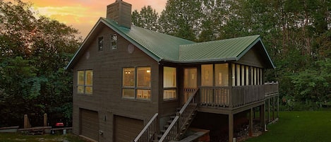 A two-story wooden cabin with a porch and a garage, surrounded by a grassy yard and trees.
