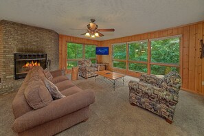 A cozy living room with a fireplace, large windows, and a ceiling fan. - Misty Cove Cabin Rental (Wears Valley)