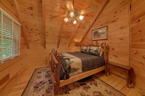 A wooden cabin bedroom with a bed, a ceiling fan, a window with blinds, and a painting on the wall.