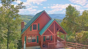 A log cabin with a green roof and a porch, surrounded by trees and mountains.
