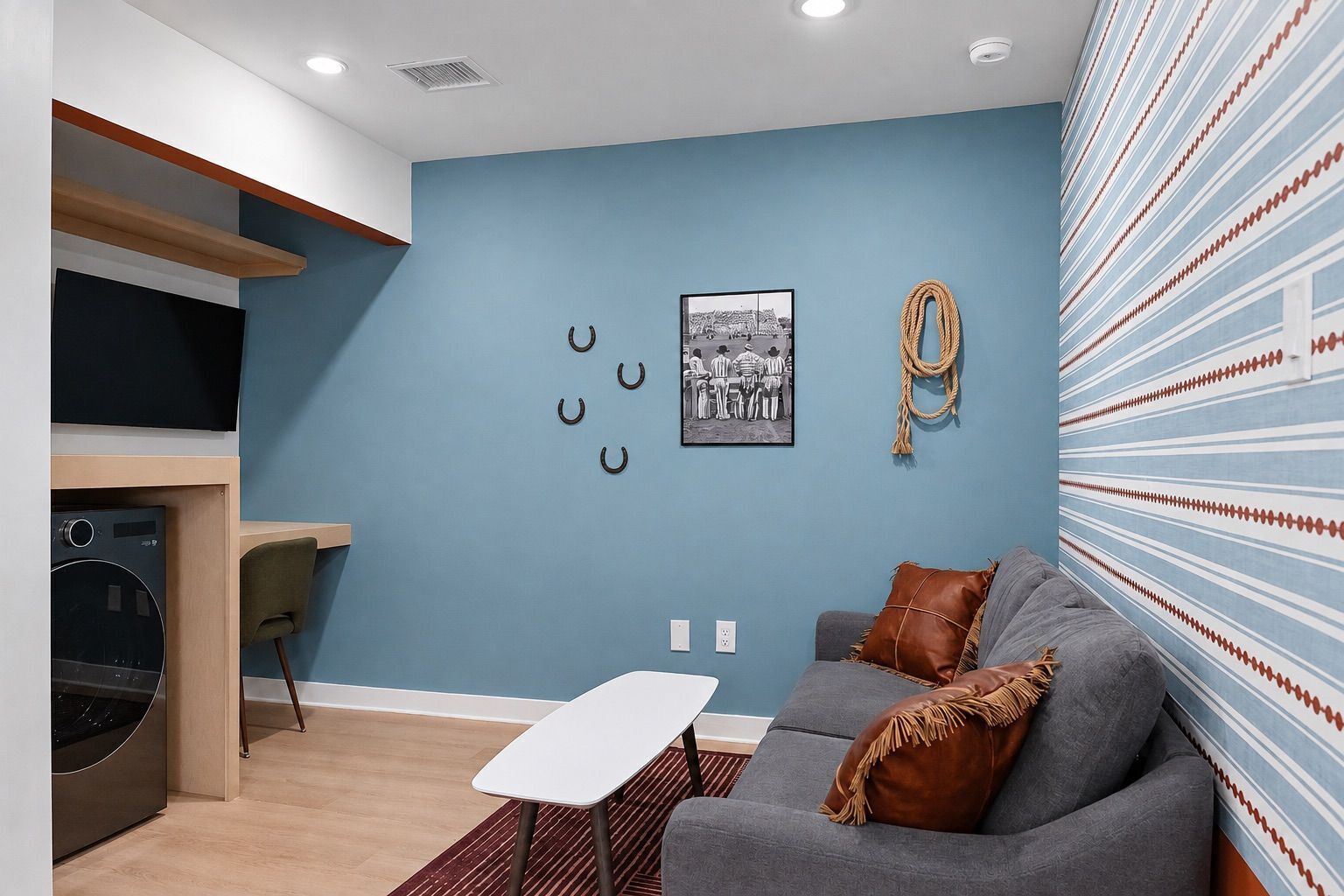 A modern living room with a blue accent wall, a grey sofa with brown cushions, a small white table, and a kitchen area with a built-in oven and microwave.