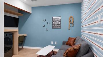 A modern living room with a blue accent wall, a grey sofa with brown cushions, a small white table, and a kitchen area with a built-in oven and microwave.