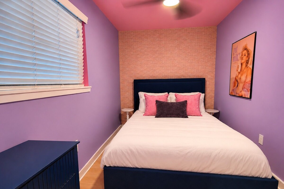 A bedroom with a bed, pillows, a nightstand, a dresser, and a window with blinds.