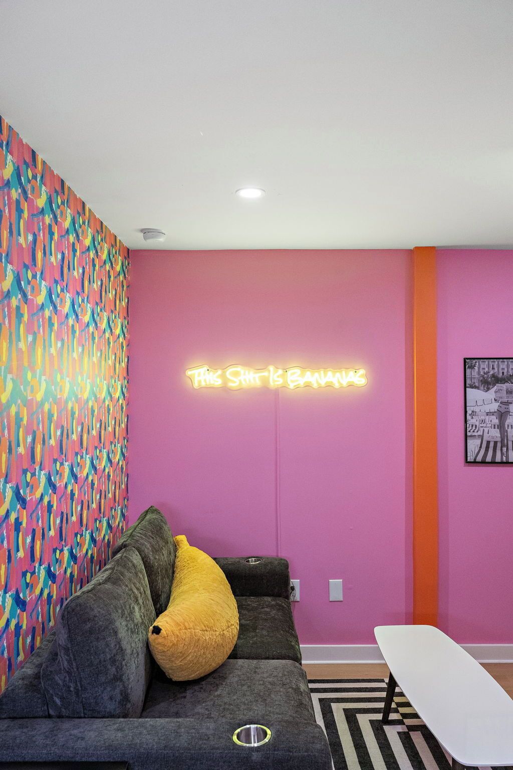 A room with a colorful abstract wall, a neon sign, a gray sofa with a yellow pillow, and a white table.