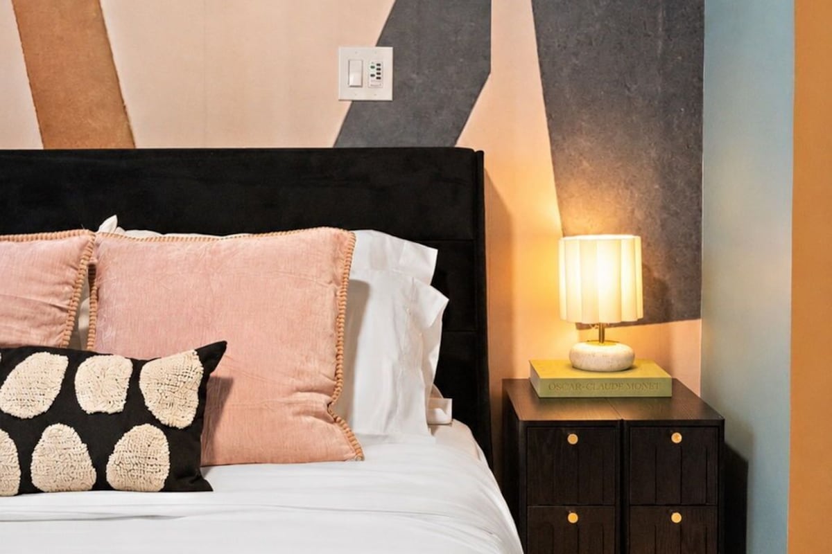 A bed with a black headboard, multiple pillows, a bedside table with a lamp, and a wall with geometric patterns.