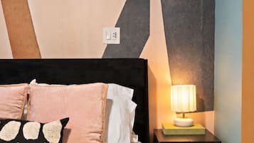 Una cama con cabecera negra, varios almohadones, una mesita de noche con lámpara y una pared con diseños geométricos.