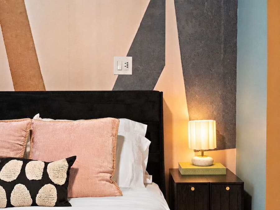 Una cama con cabecera negra, varios almohadones, una mesita de noche con lámpara y una pared con diseños geométricos.
