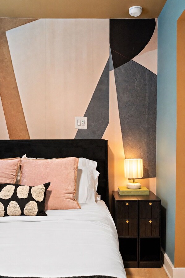 A bed with a black headboard, multiple pillows, a bedside table with a lamp, and a wall with geometric patterns.