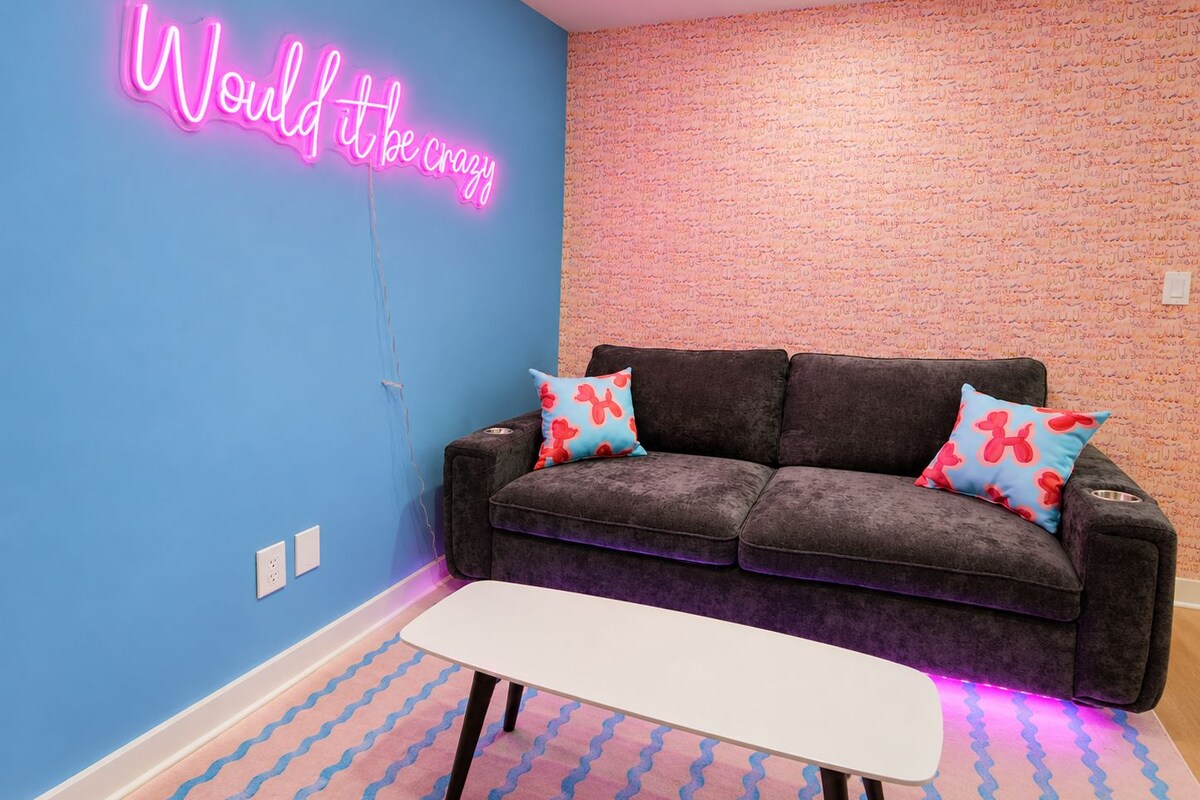 A room with a neon sign on the wall, a dark grey sofa with patterned cushions, and a white table.