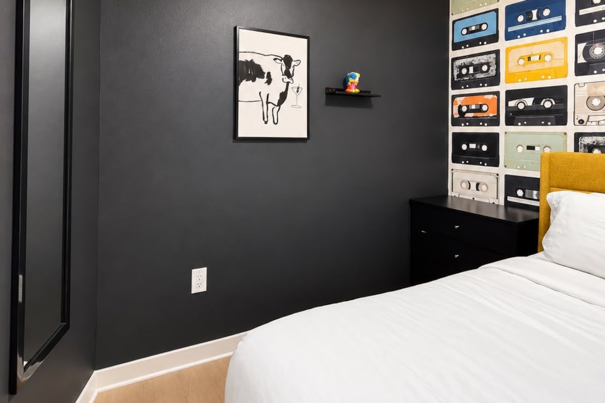 A bedroom with a bed, a ceiling fan, a wall-mounted shelf with cassette tapes, and a framed picture of a cow.