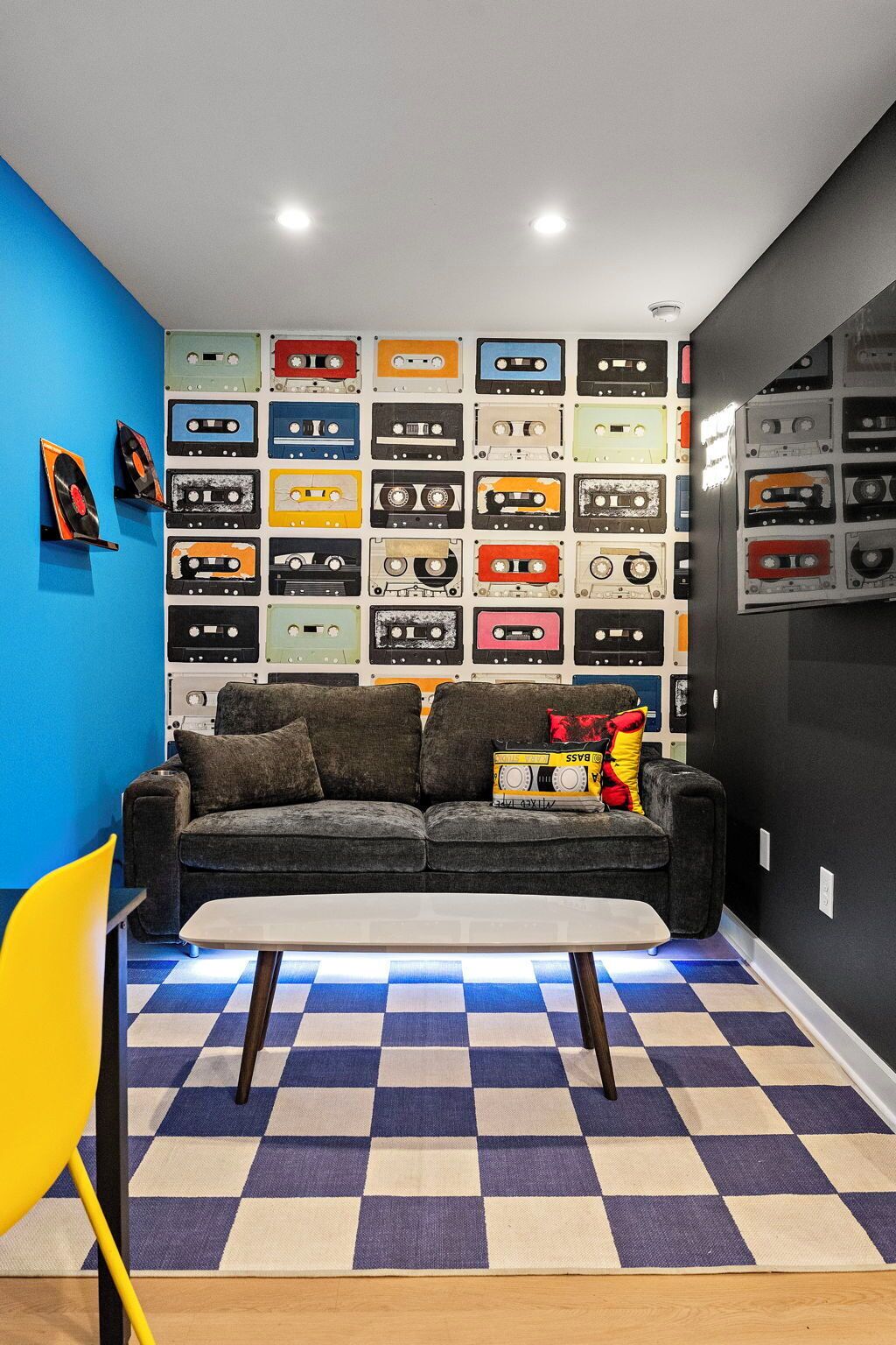 Ein Zimmer mit Schachbrettmuster-Boden, einer Couch, einem Tisch und einer Wand, die mit Kassettentapes dekoriert ist.