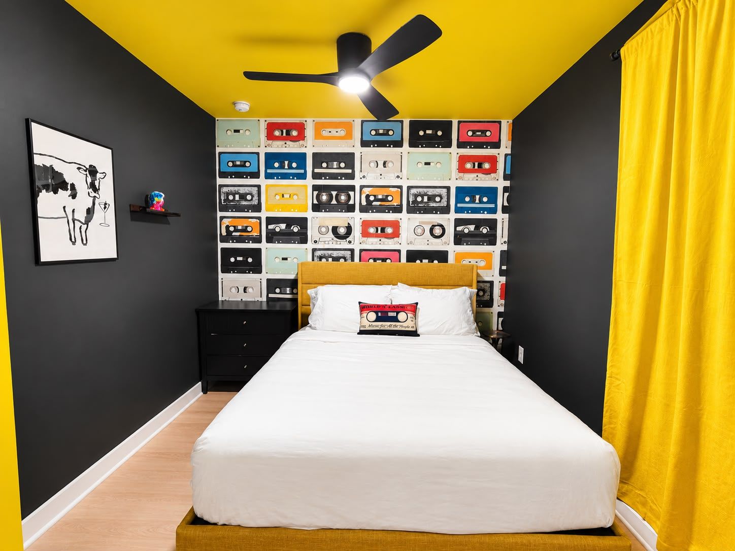 Ein Schlafzimmer mit einem Bett, einem Deckenventilator, einer Wand voller farbiger Kassetten, einem gerahmten Bild und einem Nachttisch.