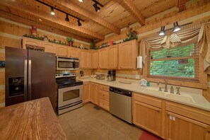 A kitchen with wooden cabinets, stainless steel appliances, and a window with a view of greenery. - Serenity Creek | Hot Tub, Resort Pool, Fireplace + Free Attraction Tickets (Wears Valley)