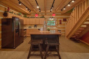 A kitchen with wooden cabinets, a stainless steel refrigerator, a wooden island with stools, and a staircase leading to an upper level. - Serenity Creek | Hot Tub, Resort Pool, Fireplace + Free Attraction Tickets (Wears Valley)