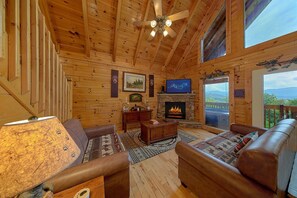 A wooden cabin interior with a fireplace, leather sofas, a television, and a scenic view through the window.