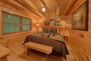 A wooden cabin bedroom with a bed, bench, and a large window with blinds.