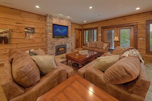 A log cabin interior with a stone fireplace, leather sofas, a coffee table, and a television mounted on the wall. - Grouse Ridge Lodge | Smoky Mtn Views, Hot Tub, Sleeps 12, Fire Pit + Free Attraction Tickets (Wears Valley)
