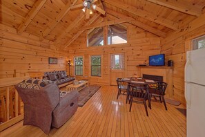 Ein Interieur einer Holzhütte mit einer Couch, einem Esstisch, Stühlen, einem Fernseher und einem Kühlschrank.