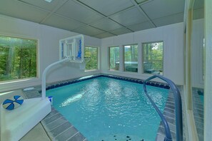 An indoor swimming pool with clear blue water, a basketball hoop, and large windows offering views of the outdoors. - Big Bad John's Cabin Rental (Wears Valley)