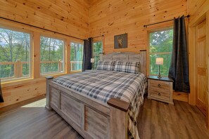 A wooden cabin bedroom with a large bed, two bedside tables, and a window overlooking a forest. - Big Bad John's Cabin Rental (Wears Valley)