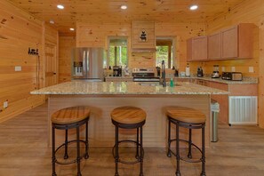 A kitchen with wooden walls, a stainless steel refrigerator, a double oven, and a granite countertop. There are three wooden stools and a stainless steel trash can. - Big Bad John's Cabin Rental (Wears Valley)