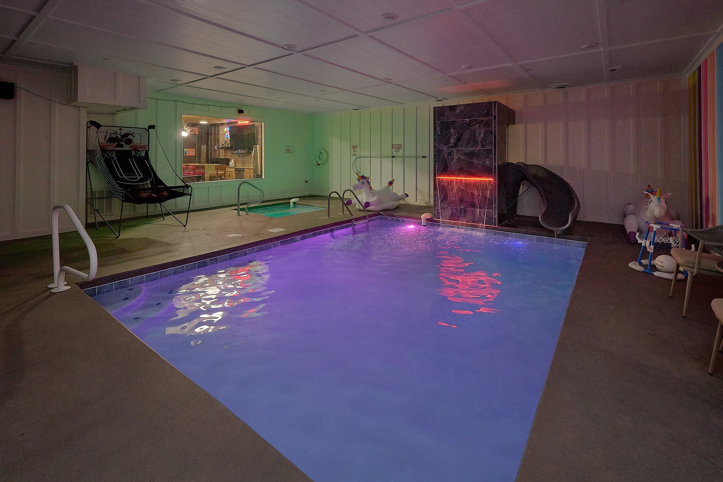 An indoor swimming pool with a basketball hoop, a lounge chair, and a small table with a unicorn decoration.