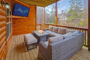 Eine Holzterrasse mit einer Couch und einem Tisch mit Feuerstelle, einem an der Wand montierten Fernseher und Blick auf Bäume.