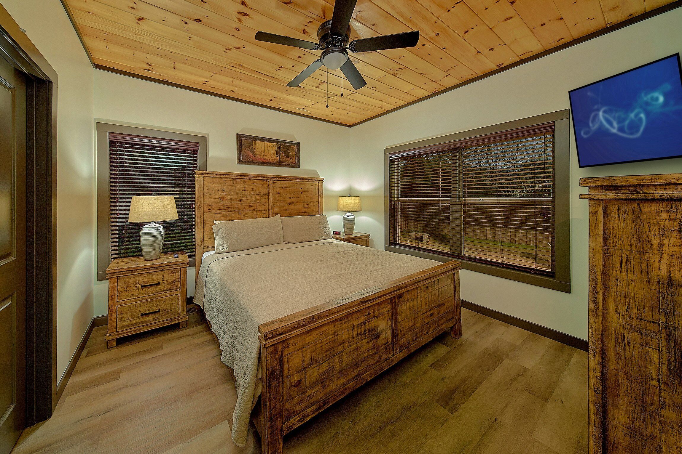Une chambre à coucher avec un lit en bois, deux tables de chevet avec des lampes, un ventilateur de plafond et une grande fenêtre avec des stores.