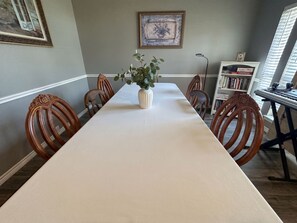 Ein Esszimmer mit einem langen Tisch, Stühlen, einer Vase mit Grünpflanzen, einem gerahmten Bild an der Wand und einem Bücherregal mit Büchern.