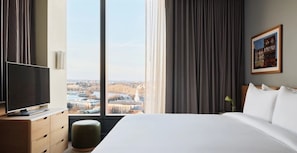 Ein Hotelzimmer mit einem großen Bett, einem Fernseher auf einer Holzkommode und Blick auf eine Stadtlandschaft durch große Fenster.
