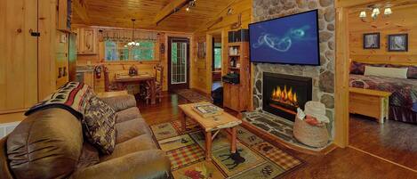 A cozy cabin interior with a stone fireplace, wooden furniture, and a television mounted on the wall.