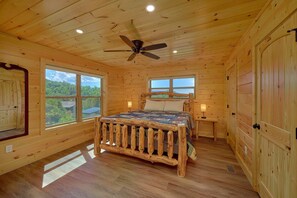 Ein Schlafzimmer in einer Holzhütte mit einem Bett, zwei Nachttischen, einem Deckenventilator und großen Fenstern.