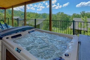 A hot tub with a control panel, surrounded by a wooden deck and a metal railing, overlooking a residential area with trees and houses.