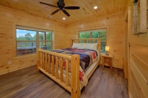 A wooden cabin bedroom with a bed, a nightstand, a ceiling fan, and large windows.