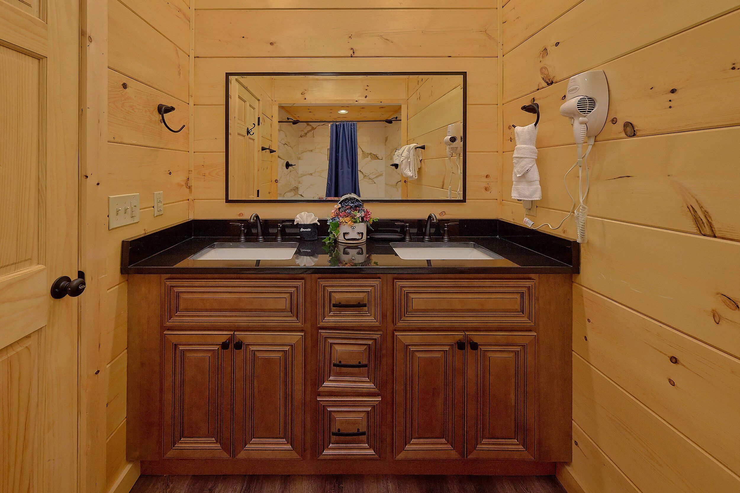 A wooden-paneled bathroom with a double sink, a mirror, and two hair dryers mounted on the wall.
