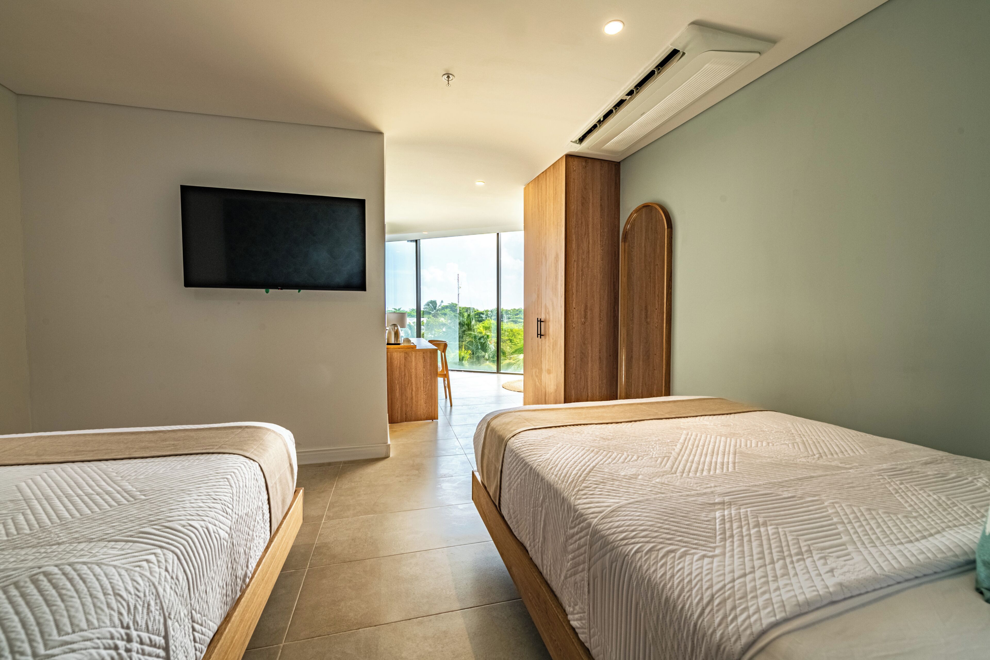 A hotel room with two beds, a flat-screen TV, a wooden wardrobe, and a view of greenery outside.