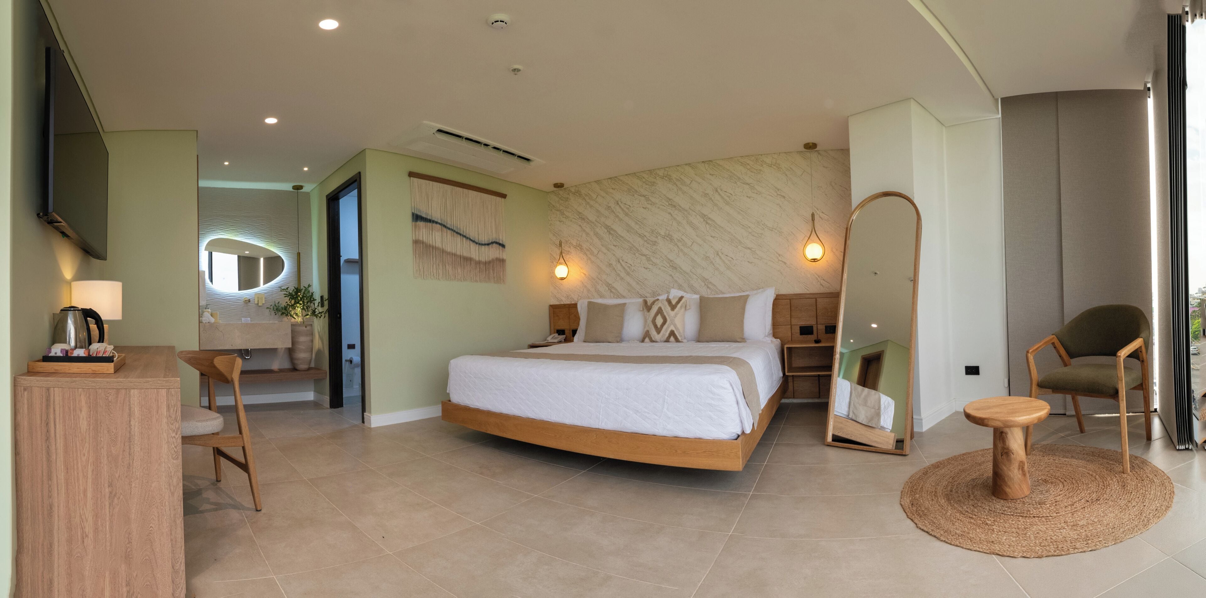 Quarto de hotel moderno com uma cama grande, um espaço de estar com uma cadeira e mesa, um espelho e vista para a cidade.