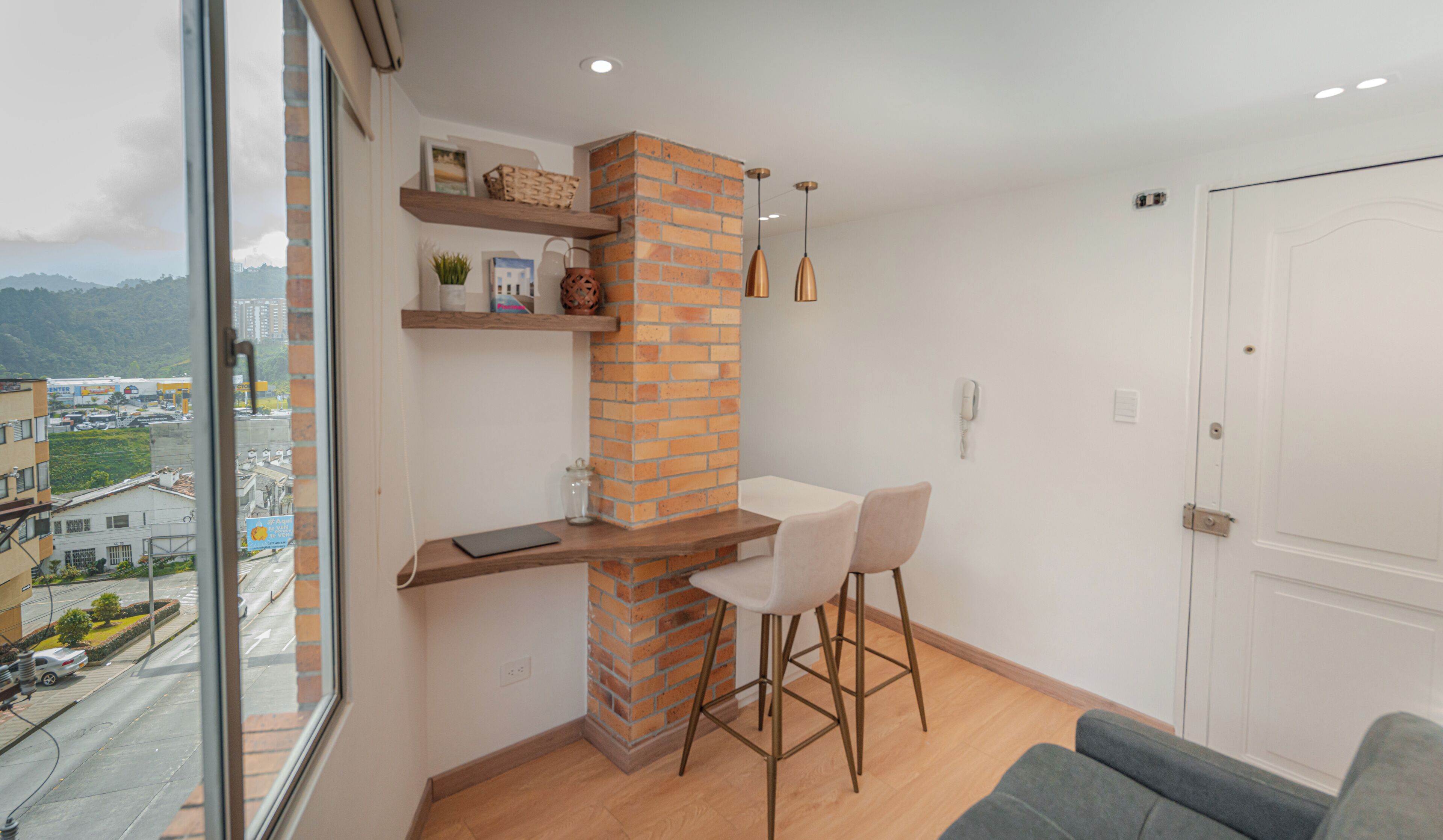 Un interior moderno con una pared de ladrillo decorativa, una silla blanca, un sofá y vistas de la ciudad a través de un ventanal grande.