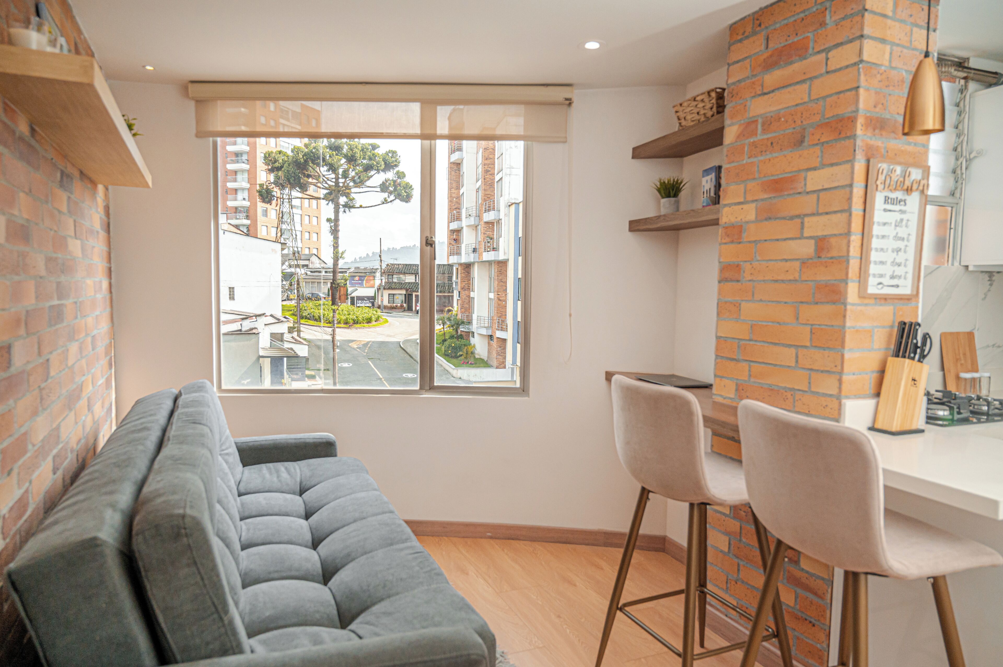 A modern kitchen with a brick accent wall, a dining area with a grey sofa and two bar stools, and a large window offering a view of the street and buildings.