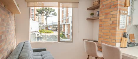 A modern kitchen with a brick accent wall, a dining area with a grey sofa and two bar stools, and a large window offering a view of the street and buildings.