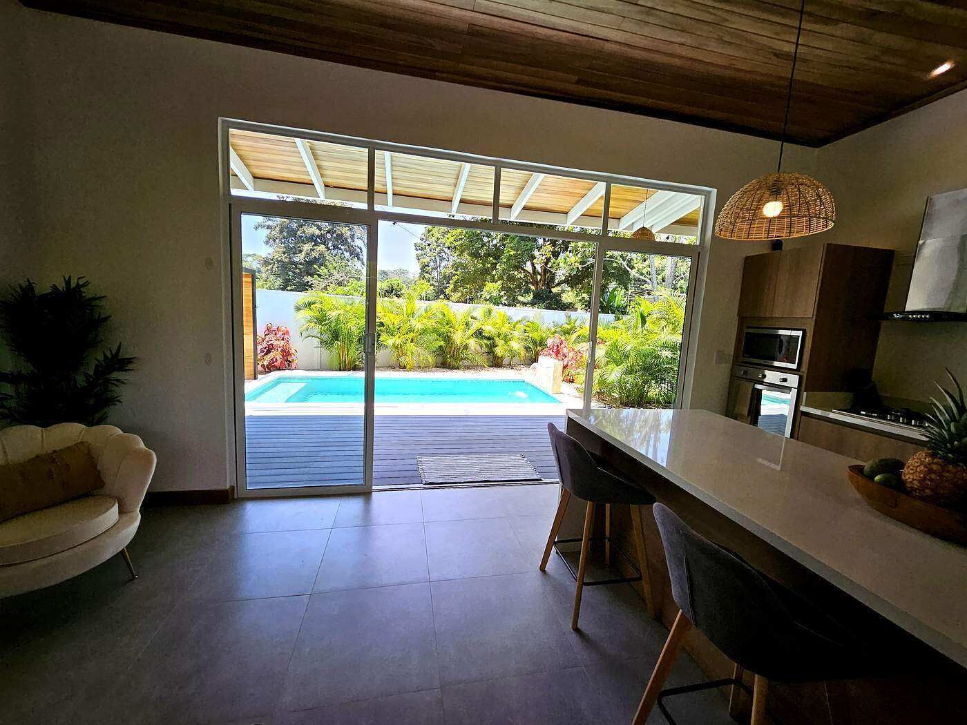 Une cuisine moderne dotée d’un grand îlot central, de portes-fenêtres coulissantes donnant sur une piscine et d’un coin repas avec un canapé.