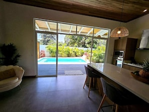 A modern kitchen with a large island, sliding glass doors leading to a pool, and a dining area with a sofa. - Papi's Place: Private Pool Villa in Playa Negra, Costa Rica (Cahuita)