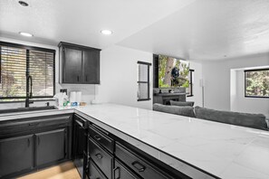 A modern kitchen with black cabinets, a white marble countertop, and a large window with blinds. - Summit Mountain Cabin (Big Bear Lake)