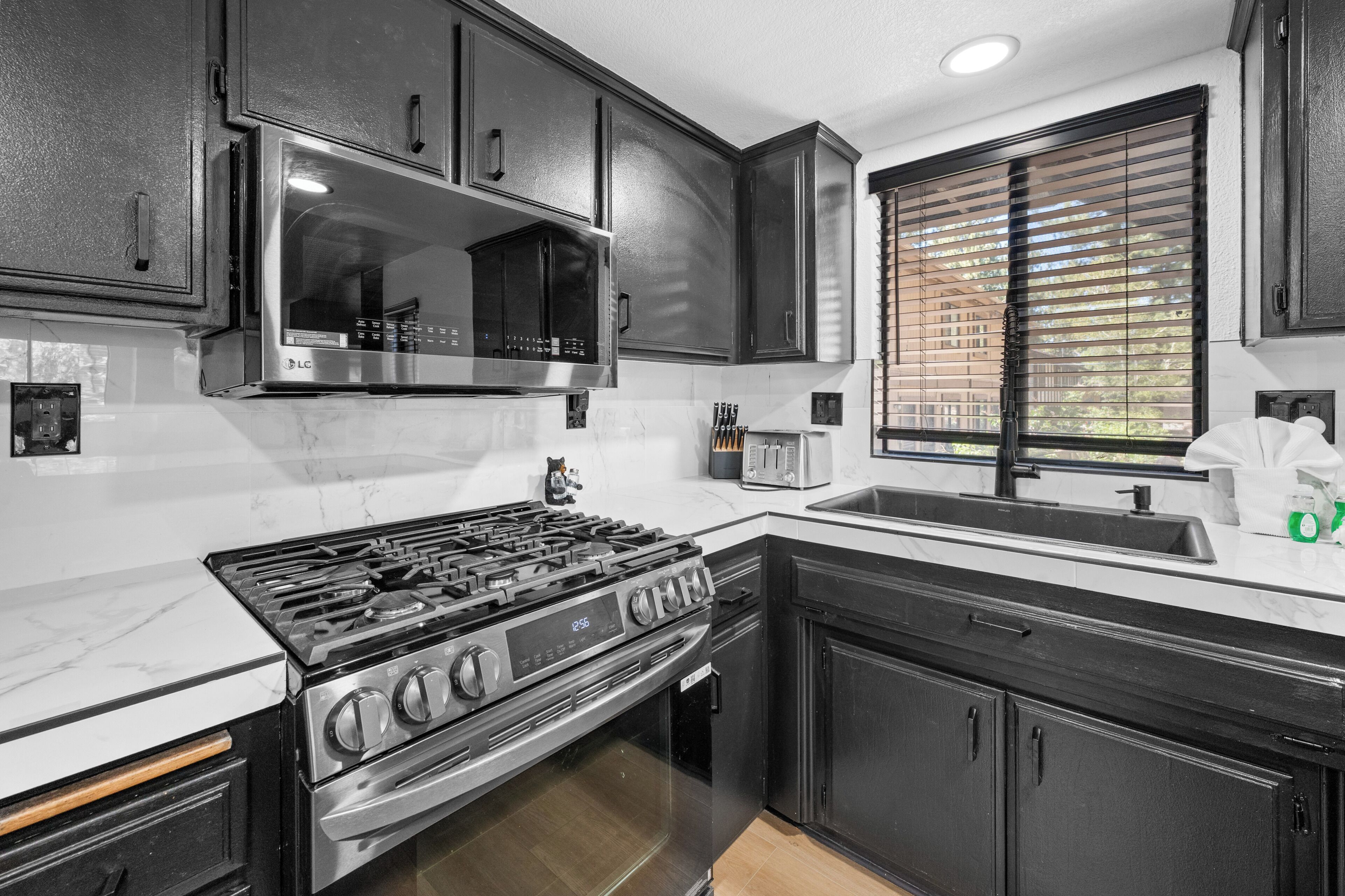 A kitchen with a stainless steel stove, black cabinets, a microwave, and a window with blinds.