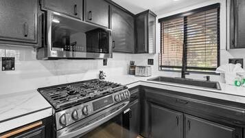 A kitchen with a stainless steel stove, black cabinets, a microwave, and a window with blinds.